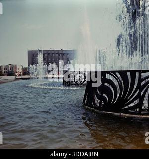 Slawjansk, Ukraine, UdSSR, 1980er Jahre Blick auf den funktionierenden Brunnen auf dem zentralen Platz der Oktoberrevolution (heute Soborna-Platz) während einer Großstadtfeier. Im Vordergrund sprühen Wasserstrahlen aus einer Struktur mit dekorativen Metallgittern nach oben. Im Hintergrund versammelt sich eine große Menschenmenge mit roten Fahnen vor dem sowjetischen Verwaltungsgebäude (Stadtrat). Dieses Archivbild fängt die festliche Atmosphäre und die urbane Landschaft der Donbass-Stadt ein. Stockfoto