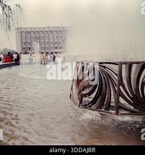 Slawjansk, Ukraine, UdSSR, 1980er Jahre Blick auf den funktionierenden Brunnen auf dem zentralen Platz der Oktoberrevolution (heute Soborna-Platz) während einer Großstadtfeier. Im Vordergrund sprühen Wasserstrahlen aus einer Struktur mit dekorativen Metallgittern nach oben. Im Hintergrund versammelt sich eine große Menschenmenge mit roten Fahnen vor dem sowjetischen Verwaltungsgebäude (Stadtrat). Dieses Archivbild fängt die festliche Atmosphäre und die urbane Landschaft der Donbass-Stadt ein. Stockfoto