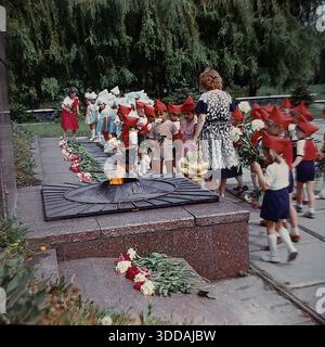 Slawjansk, Ukraine, UdSSR, 1980er Jahre Eine Gruppe von Kindergartenkindern in festlicher Uniform (rote Spitzhüte, die Budenovkas ähneln, rote Hemden, blaue Shorts) legt Blumen an der Gedenkstätte für die Ewige Flamme im Lenin Park (heute Shelkovichny Park). Lehrer begleiten die Kinder während einer feierlichen patriotischen Zeremonie zu Ehren der Veteranen des Zweiten Weltkriegs. Eine weitere Gruppe von Kindern in blau-weißen Kostümen wartet im Hintergrund. Dieses Archivbild fängt die Tradition der sowjetischen patriotischen Erziehung ein. Stockfoto
