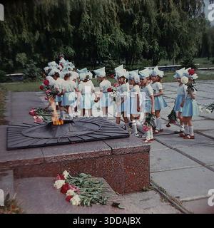 Slawjansk, Ukraine, UdSSR, 1980er Jahre Eine feierliche Prozession von Kindergartenkindern in festlichen blau-weißen Kostümen mit spitzen Hüten am Denkmal der Ewigen Flamme im Lenin Park (heute Shelkovichny Park). Die Jungen und Mädchen halten frische Blumen (rote und weiße Pfingstrosen), während sie in einer Linie an dem brennenden sternförmigen Gasbrenner vorbeilaufen. Eine Lehrerin überwacht die patriotische Zeremonie zu Ehren der Veteranen des Zweiten Weltkriegs. Dieses Archivbild fängt die Tradition der sowjetischen Kindererziehung und des Gedenkens ein. Stockfoto