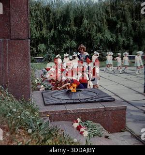Slawjansk, Ukraine, UdSSR, 1980er Jahre Eine Gruppe von Kindergartenkindern in festlicher Uniform (rote Spitzhüte, die Budenovkas ähneln, rote Hemden, blaue Shorts) legt Blumen an der Gedenkstätte für die Ewige Flamme im Lenin Park (heute Shelkovichny Park). Lehrer begleiten die Kinder während einer feierlichen patriotischen Zeremonie zu Ehren der Veteranen des Zweiten Weltkriegs. Eine weitere Gruppe von Kindern in blau-weißen Kostümen wartet im Hintergrund. Dieses Archivbild fängt die Tradition der sowjetischen patriotischen Erziehung ein. Stockfoto