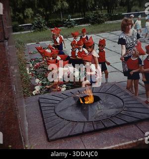 Slawjansk, Ukraine, UdSSR, 1980er Jahre Eine Gruppe von Kindergartenkindern in festlicher Uniform (rote Spitzhüte, die Budenovkas ähneln, rote Hemden, blaue Shorts) legt Blumen an der Gedenkstätte für die Ewige Flamme im Lenin Park (heute Shelkovichny Park). Lehrer begleiten die Kinder während einer feierlichen patriotischen Zeremonie zu Ehren der Veteranen des Zweiten Weltkriegs. Eine weitere Gruppe von Kindern in blau-weißen Kostümen wartet im Hintergrund. Dieses Archivbild fängt die Tradition der sowjetischen patriotischen Erziehung ein. Stockfoto