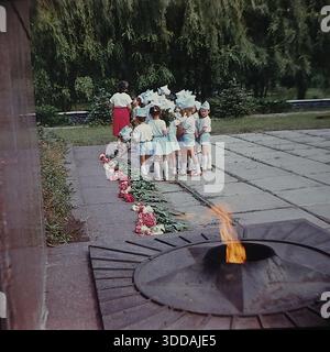 Slawjansk, Ukraine, UdSSR, 1980er Jahre Eine Gruppe von Kindergartenkindern in festlichen weißen und hellblauen Kostümen mit passenden Hüten legt Blumen am Denkmal für die Ewige Flamme im Lenin Park (heute Shelkovichny Park). Eine Lehrerin in einem roten Rock leitet die Zeremonie. Die Kinder stehen in einer Schlange neben dem Granitsternbrenner mit brennendem Feuer. Rote Nelken und Pfingstrosen sind auf den Stein gelegt. Dieses Archivbild fängt die Tradition der patriotischen Erziehung in der Sowjetunion ein. Stockfoto