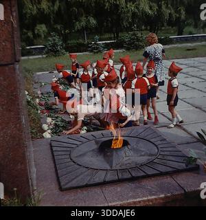 Slawjansk, Ukraine, UdSSR, 1980er Jahre Eine Gruppe von Kindergartenkindern in festlicher Uniform (rote Spitzhüte, die Budenovkas ähneln, rote Hemden, blaue Shorts) legt Blumen an der Gedenkstätte für die Ewige Flamme im Lenin Park (heute Shelkovichny Park). Lehrer begleiten die Kinder während einer feierlichen patriotischen Zeremonie zu Ehren der Veteranen des Zweiten Weltkriegs. Eine weitere Gruppe von Kindern in blau-weißen Kostümen wartet im Hintergrund. Dieses Archivbild fängt die Tradition der sowjetischen patriotischen Erziehung ein. Stockfoto