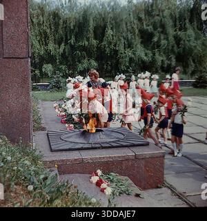 Slawjansk, Ukraine, UdSSR, 1980er Jahre Eine Gruppe von Kindergartenkindern in festlicher Uniform (rote Spitzhüte, die Budenovkas ähneln, rote Hemden, blaue Shorts) legt Blumen an der Gedenkstätte für die Ewige Flamme im Lenin Park (heute Shelkovichny Park). Lehrer begleiten die Kinder während einer feierlichen patriotischen Zeremonie zu Ehren der Veteranen des Zweiten Weltkriegs. Eine weitere Gruppe von Kindern in blau-weißen Kostümen wartet im Hintergrund. Dieses Archivbild fängt die Tradition der sowjetischen patriotischen Erziehung ein. Stockfoto