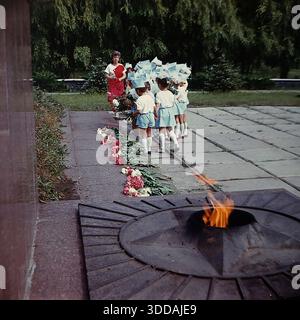 Slawjansk, Ukraine, UdSSR, 1980er Jahre Eine Gruppe von Kindergartenkindern in festlichen weißen und hellblauen Kostümen mit passenden Hüten legt Blumen am Denkmal für die Ewige Flamme im Lenin Park (heute Shelkovichny Park). Eine Lehrerin in einem roten Rock leitet die Zeremonie. Die Kinder stehen in einer Schlange neben dem Granitsternbrenner mit brennendem Feuer. Rote Nelken und Pfingstrosen sind auf den Stein gelegt. Dieses Archivbild fängt die Tradition der patriotischen Erziehung in der Sowjetunion ein. Stockfoto