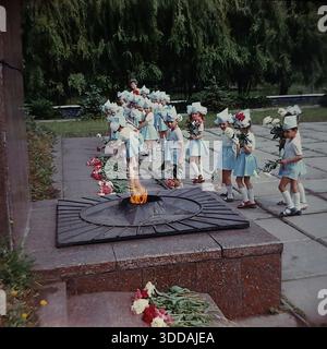 Slawjansk, Ukraine, UdSSR, 1980er Jahre Eine feierliche Prozession von Kindergartenkindern in festlichen blau-weißen Kostümen mit spitzen Hüten am Denkmal der Ewigen Flamme im Lenin Park (heute Shelkovichny Park). Die Jungen und Mädchen halten frische Blumen (rote und weiße Pfingstrosen), während sie in einer Linie an dem brennenden sternförmigen Gasbrenner vorbeilaufen. Eine Lehrerin überwacht die patriotische Zeremonie zu Ehren der Veteranen des Zweiten Weltkriegs. Dieses Archivbild fängt die Tradition der sowjetischen Kindererziehung und des Gedenkens ein. Stockfoto