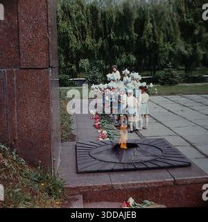 Slawjansk, Ukraine, UdSSR, 1980er Jahre Eine Gruppe von Kindergartenkindern in festlichen weißen und hellblauen Kostümen mit passenden Hüten legt Blumen am Denkmal für die Ewige Flamme im Lenin Park (heute Shelkovichny Park). Eine Lehrerin in einem roten Rock leitet die Zeremonie. Die Kinder stehen in einer Schlange neben dem Granitsternbrenner mit brennendem Feuer. Rote Nelken und Pfingstrosen sind auf den Stein gelegt. Dieses Archivbild fängt die Tradition der patriotischen Erziehung in der Sowjetunion ein. Stockfoto