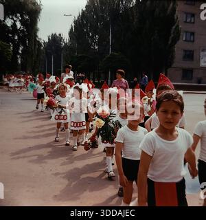 Slawjansk, Ukraine, UdSSR, 1980er Jahre Eine festliche Prozession von Kindergartenkindern, die während einer Feiertagsfeier auf einer Stadtallee spazieren. Die Spalte enthält verschiedene Gruppen in Kostümen: Mädchen in weißen Kleidern mit roten Ringmustern, Jungen in Sportuniformen und Kinder in roten Spitzhüten (budenovkas) im Hintergrund. Viele Kinder tragen frische Pfingstrosen und Ballons. Lehrer überwachen die Parade. Dieses Archivbild zeigt das Ausmaß und die Atmosphäre der sowjetischen patriotischen Feiertage. Stockfoto