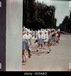 Slawjansk, Ukraine, UdSSR, 1980er Jahre Ein offenes Bild einer Kindergartenprozession, die durch den Lenin Park (heute Shelkovichny Park) geht. Die Kinder tragen dazu passende festliche blau-weiße Kostüme mit gestreiften Shorts und spitzen Hüten. Sie halten Hände und tragen große Sträuße rosa Pfingstrosen. Eine Lehrerin im roten Rock folgt der Gruppe. Das Bild wird von einer weißen Säule im Vordergrund eingerahmt. Dieses Archivfoto zeigt die Unschuld und patriotische Traditionen der sowjetischen Kindheit. Stockfoto