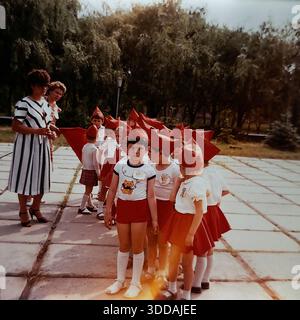 Slawjansk, Ukraine, UdSSR, 1980er Jahre Eine Gruppe von Kindergartenkindern in festlichen rot-weißen Uniformen bereitet sich auf eine patriotische Vorstellung im Stadtpark vor. Die zentrale Figur ist ein ernsthafter Junge mit einem roten Spitzhut im Budenovka-Stil und einem T-Shirt mit Katzenmotiv. Andere Kinder halten kleine rote Fahnen. Zwei Lehrerinnen, eine in einem stilvollen schwarz-weiß gestreiften Kleid, beaufsichtigen die Kinder. Dieses Archivfoto fängt die Atmosphäre der sowjetischen Kindheit und Feiertage ein. Stockfoto