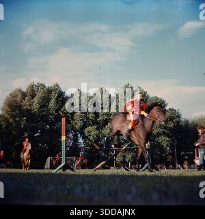 Slawjansk, Ukraine, UdSSR, 1980er Jahre Eine dynamische Action-Aufnahme eines Reiters aus der Bezirksreitersportschule, der eine Springübung im Khimik-Stadion durchführt. Der Athlet trägt ein festliches rotes Husaren-Kostüm mit goldener Flechtung. Das Pferd springt über eine Holzbarriere auf dem grünen Rasen des Fußballfeldes. Im Hintergrund sind Fußballtorpfosten und andere Fahrer zu sehen. Dieses Archivfoto zeigt ein Training oder eine öffentliche Demonstration des sowjetischen Reitsports. Stockfoto