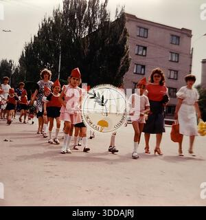 Slawjansk, Ukraine, UdSSR, 1980er Jahre Eine festliche Straßenprozession zum Internationalen Kindertag (1. Juni). Kinder aus dem Kindergarten „Oduvanchik“ (Löwenzahn) marschieren mit einem großen Emblem, das eine Taube des Friedens, eine Kugel und eine Löwenzahn-Blume zeigt. Die Kinder tragen Kostüme mit roten spitzen Hüten. Lehrer und Eltern begleiten die Gruppe. Im Hintergrund ist ein typisches sowjetisches Wohngebäude zu sehen. Dieses Archivfoto zeigt die pazifistische Erziehung in der UdSSR. Stockfoto