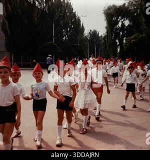 Slawjansk, Ukraine, UdSSR, 1980er Jahre Eine große Gruppe von Kindergartenkindern marschiert in einer festlichen Prozession zum Internationalen Kindertag (1. Juni). Die Kinder tragen Kostüme mit roten spitzen Hüten (Budenovka-Stil), weißen Hemden und Shorts. Ein Mädchen in einem weißen Spitzenkleid geht in die Mitte. Sie halten Hände und gehen eine Straße entlang, die von hohen Pappelbäumen gesäumt ist. Ein sowjetischer Polizist in blauer Uniform überwacht die Sicherheit. Dieses Archivfoto zeigt eine glückliche sowjetische Kindheit. Stockfoto