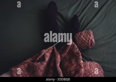 Melierter brauner Pullover mit Stricknadeln und Frauenbeinen in Samtsocken auf dunkelgrünem Hintergrund. Stricknadeln und Kugeln aus warmem, meliertem Garn. Stockfoto
