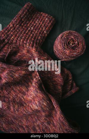 Melierter brauner Pullover mit Stricknadeln und Frauenbeinen in Samtsocken auf dunkelgrünem Hintergrund. Stricknadeln und Kugeln aus warmem, meliertem Garn. Stockfoto