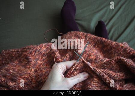 Melierter brauner Pullover mit Stricknadeln und Frauenbeinen in Samtsocken auf dunkelgrünem Hintergrund. Stricknadeln und Kugeln aus warmem, meliertem Garn. Stockfoto