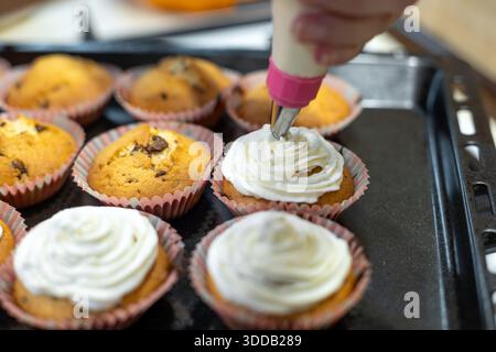 Cupcakes mit Zuckerguss auf Backblech dekorieren. Nahaufnahme von Handputzen cremeweißem Zuckerguss auf frisch gebackenen Cupcakes in Papiereinlagen, bereit für Stockfoto