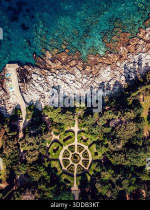 Blick aus der Vogelperspektive auf das türkisfarbene Meer, das die zerklüfteten, weißen Felsen küsst, während ein sorgfältig gestalteter Garten inmitten üppiger Vegetation in Dubrovnik, Dubrovnik liegt Stockfoto