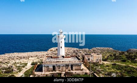 Weißer Leuchtturm mit sichtbaren Schäden steht an der felsigen Küste in der Nähe eines verlassenen Gebäudes, mit Blick auf das ruhige Meer und die zerklüftete Landschaft unter blauem Himmel Stockfoto
