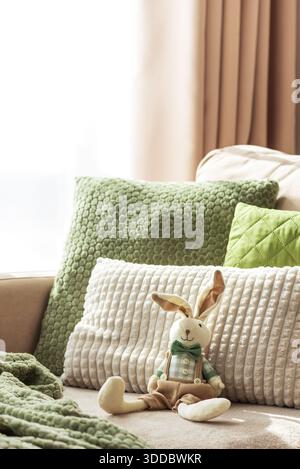 Gemütliches Sofa mit weicher Osterhase-Puppe und strukturierten Kissen im Sonnenlicht Stockfoto