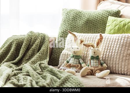 Gemütliches Sofa mit weichen Osterhasen-Puppen und strukturierten Kissen im Sonnenlicht Stockfoto
