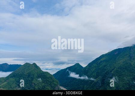 Weiche Wolken schweben über steilen, bewaldeten Bergen in der Nähe von Nong Khiaw, Laos. Die Landschaft bietet üppige grüne Hänge und einen sich windenden Fluss unter einem breiten, bewölkten Himmel. Stockfoto