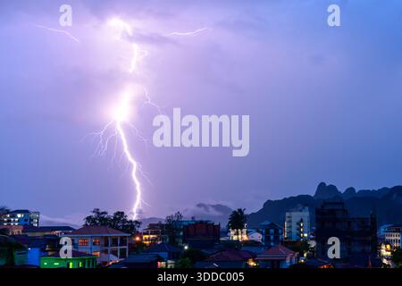 Ein dramatischer Blitz beleuchtet den Abendhimmel über den Dächern von Vang Vieng, Laos, mit Silhouetten von Kalksteinbergen im Hintergrund. Die stürmische Atmosphäre schafft einen lebhaften Kontrast zwischen dem elektrischen Licht und der ruhigen Stadt darunter. Stockfoto