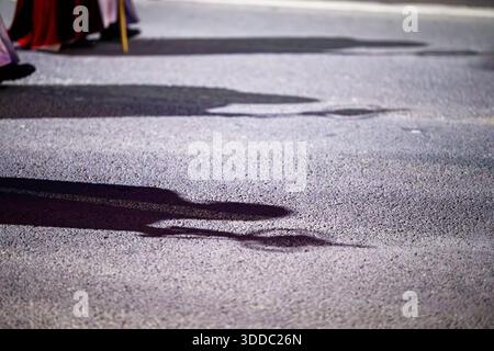 Eine niederwinklige, künstlerische Aufnahme, die sich auf die langen, dramatischen Schatten konzentriert, die die Pönitenten und ihre Notenblätter während des Nachts auf der Asphaltstraße hinterlassen haben Stockfoto