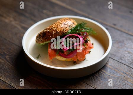 Ein Sandwich mit Zwiebeln und Lachs auf einem weißen Teller. Das Sandwich wird auf einen Holztisch gestellt Stockfoto
