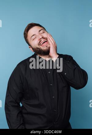 Mann in schwarzem Hemd lacht mit Hand auf Wange vor blauem Hintergrund. Stockfoto
