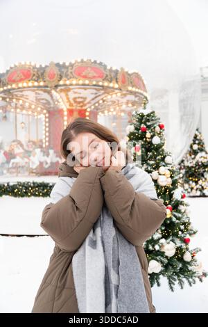 Frau steht neben geschmückten Weihnachtsbäumen und glühendem Karussell in einer festlichen Installation mit Nussknacker-Thema, umgeben von Schnee und warmen Lichtern Stockfoto