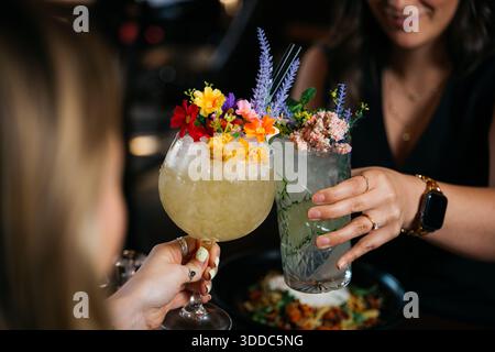 Zwei Frauen halten ihre Drinks hoch, von denen eine eine eine Blumenkarnitur hat. Die Szene spielt in einem Restaurant, und die Frauen feiern oder genießen Stockfoto