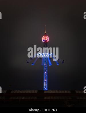Beleuchteter Schaukelturm für den Vergnügungspark bei Nacht mit blauen und roten Lichtern vor einem dunklen bewölkten Himmel. Stockfoto