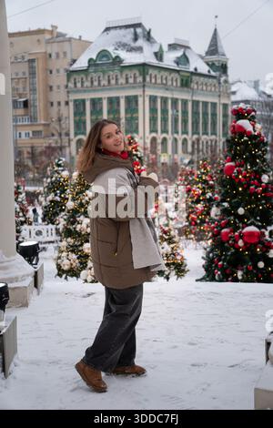 Smiling woman stands among decorated Christmas trees with snow and warm festive lights, creating a cozy urban holiday atmosphere Stockfoto