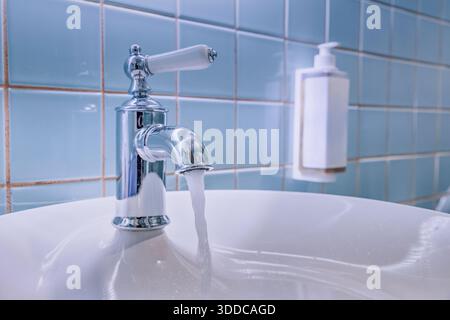 Fließendes Leitungswasser strömt in ein sauberes weißes Keramikbecken mit einem Vintage-Chromhahn gegen die blau geflieste Badezimmerwand Stockfoto