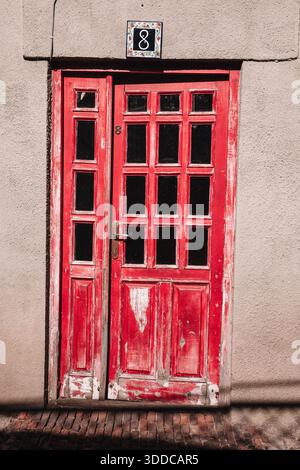 Abblätternde rote Farbe auf einer alten Holztür mit Glaspaneelen und einer Keramikfliese Nr. 8 an einer strukturierten Wand Stockfoto
