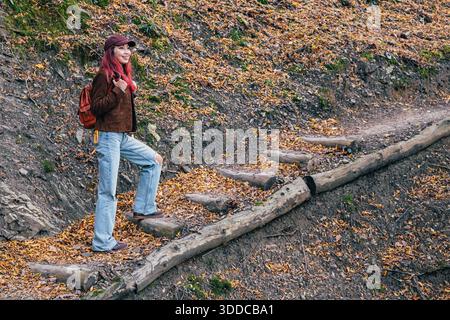 Junge Frau mit Rucksack klettert auf einem lebhaften Herbstwaldpfad auf rustikalen Holztreppen, lächelt zwischen gefallenen Blättern und Laub Stockfoto