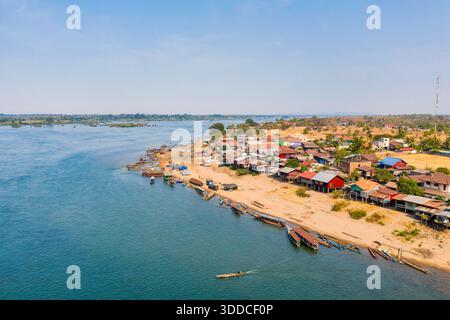 Aus der Vogelperspektive eines lebendigen Dorfes am Fluss mit Pfahlhäusern und langen Holzbooten, die eine Sandbank auf dem Mekong-Fluss in der Region 4000 Islands im Süden von Laos säumen. Helles Tageslicht offenbart ruhiges blaues Wasser, rustikale Architektur und verstreutes Grün in einer geräumigen, ruhigen Landschaft. Stockfoto