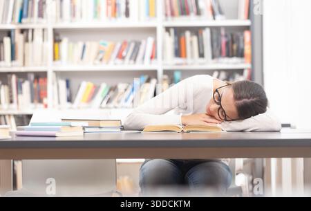 Eine junge Studentin schläft in einer Bibliothek und ruht sich auf einem Lehrbuch aus. Konzept von akademischem Stress, Erschöpfung, Studienüberlastung und Student l Stockfoto
