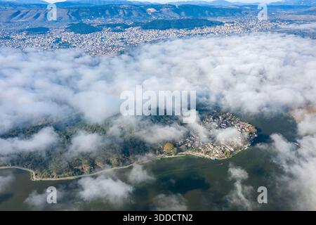 Aus der Vogelperspektive ist der schwirrende Morgennebel, der teilweise das Inseldorf am Ioannina-See enthüllt, wobei die Stadt und die Pindus-Berge eine ferne Kulisse in Epirus, Griechenland, bilden. Das Bild zeigt dichten Wald, Häuserblocks und ruhiges Wasser im See bei weichem Tageslicht. Stockfoto
