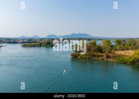 Ein kleines Boot fährt über den breiten Mekong-Fluss in der 4000-Inseln-Region im Süden von Laos. Entfernte Dörfer, üppige Flüsse und trübe blaue Hügel schaffen eine ruhige Flusslandschaft bei klarem Tageslicht. Stockfoto