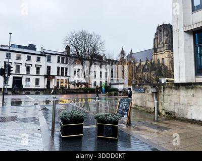 Manchester, Großbritannien - 06. Dezember 2025: Feuchte Stadtstraße in Manchester mit einer historischen Kirche, Geschäften und Fußgängern. Stockfoto