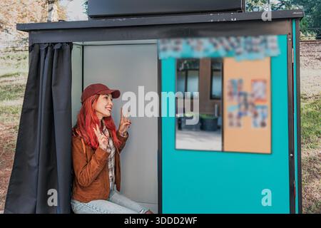 Lächelnde junge Frau mit rotem Haar, die in einer Fotokabine sitzt und den Moment genießt Stockfoto