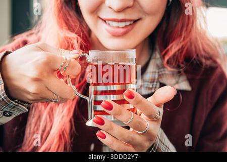 Frau mit roter Maniküre, die eine Tasse heißen Tee oder Aufguss mit einem fröhlichen, entspannten Lächeln genießt Stockfoto