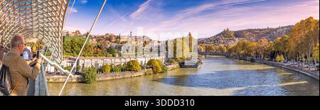 31. Oktober 2025, Tiflis, Georgien: Touristen erkunden moderne Architektur auf der Friedensbrücke mit Blick auf den Fluss Kura und die historische Stadtlandschaft von Tiflis Stockfoto