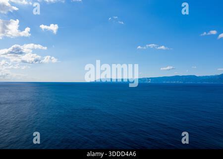 Weite Ausdehnung des tiefblauen Mittelmeers, die sich in Richtung einer trüben bergigen Küste unter einem hellen Himmel mit verstreuten Wolken in der Nähe von Orosei, Sardinien erstreckt. Stockfoto