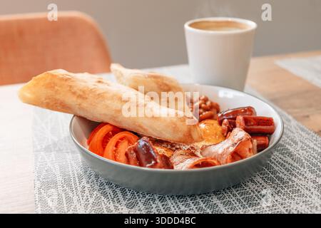 Traditioneller englischer Frühstücksteller mit Wurst, Speck, gebackenen Bohnen, Eiern und frischem Brot, serviert mit Kaffee Stockfoto