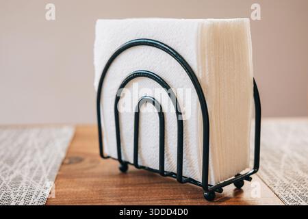 Weiße Papierservietten, ordentlich in einem minimalistischen Metallhalter angeordnet, sitzen auf einem hölzernen Esstisch Stockfoto