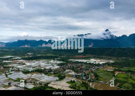 Ausgedehnte überflutete Reisfelder erstrecken sich über die Landschaft in der Nähe von Vang Vieng, Laos, mit einem Dorf, das unter dramatischen Kalksteinbergen und tief hängenden Wolken liegt. Der bewölkte Himmel und das reflektierende Wasser schaffen eine ruhige ländliche Landschaft. Stockfoto